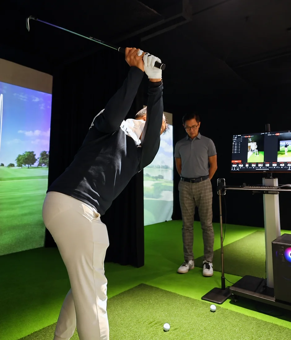 An instructor in a grey polo observes a golfer at the top of his backswing beside the replay screen